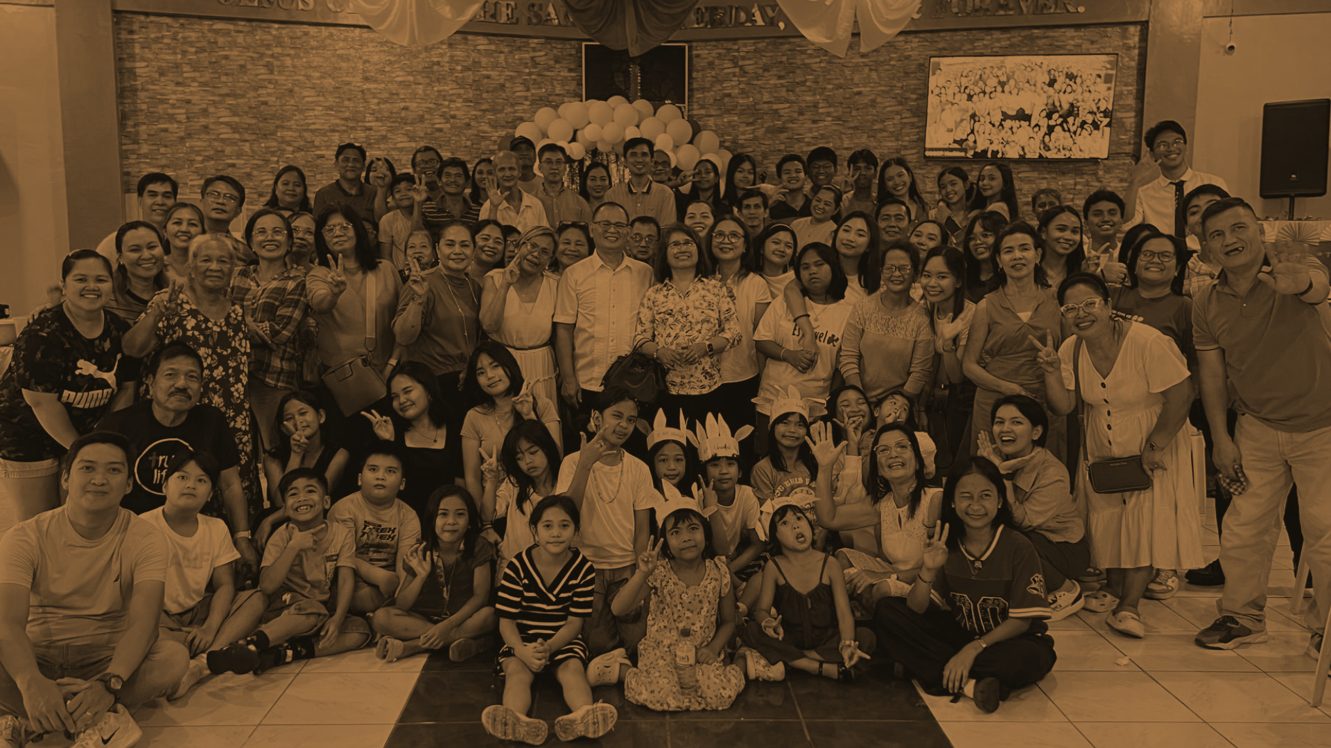 Church of God of Prophecy congregation gathered together in celebration, colorful group photo inside the church hall with blue walls, balloons, and children in Pikachu hats